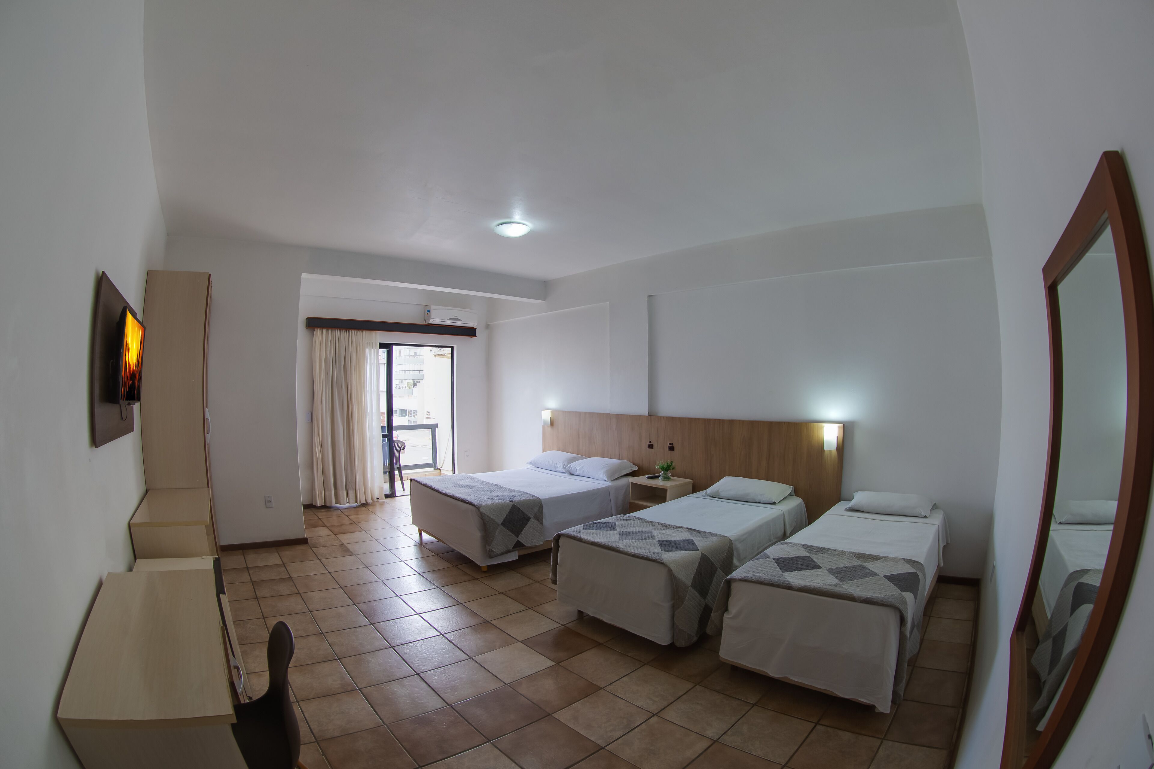 A hotel room with three beds, a desk, and a chair. There is a window with curtains, a door, and a mirror on the wall.