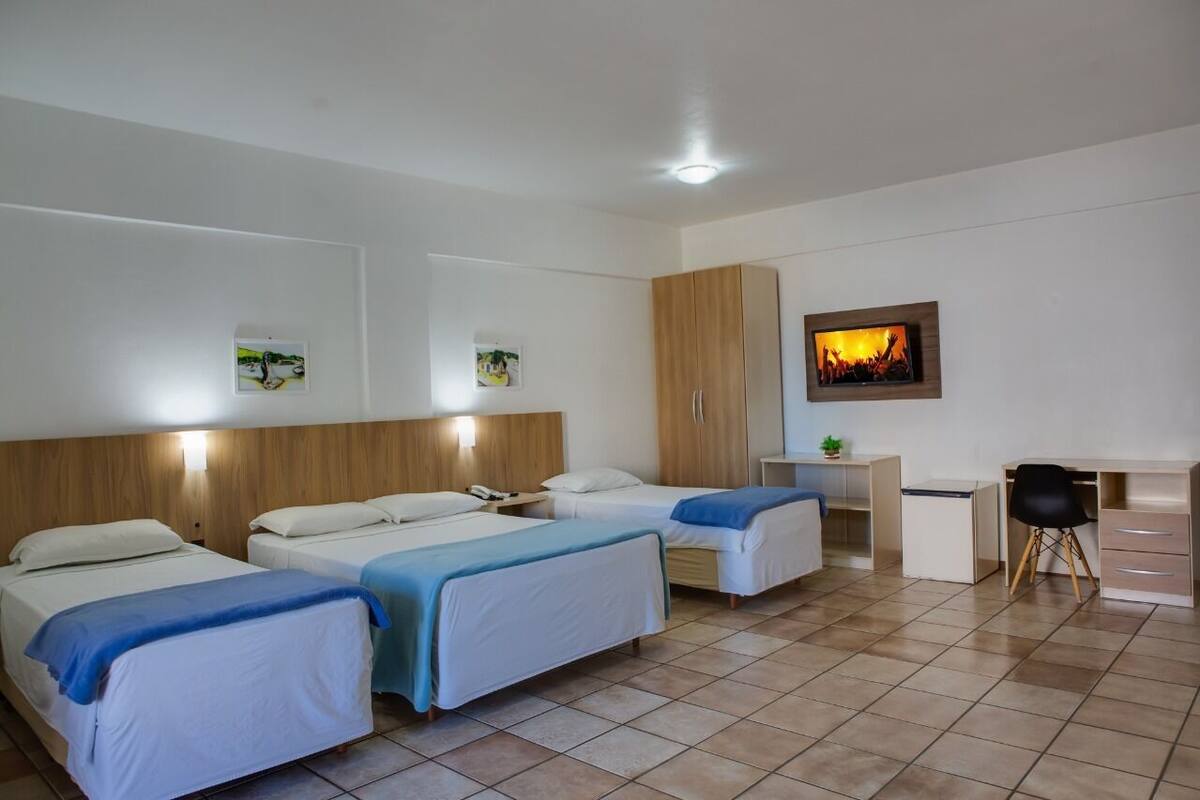 Quarto de hotel com três camas de solteiro, um guarda-roupa de madeira, uma escrivaninha com cadeira e uma televisão na parede.