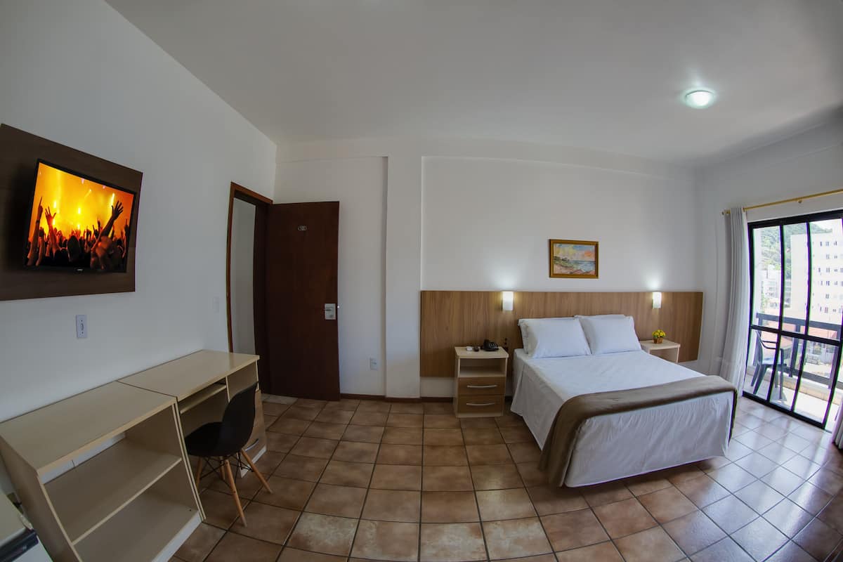 Quarto de hotel com cama, escrivaninha, cadeira e televisão na parede.