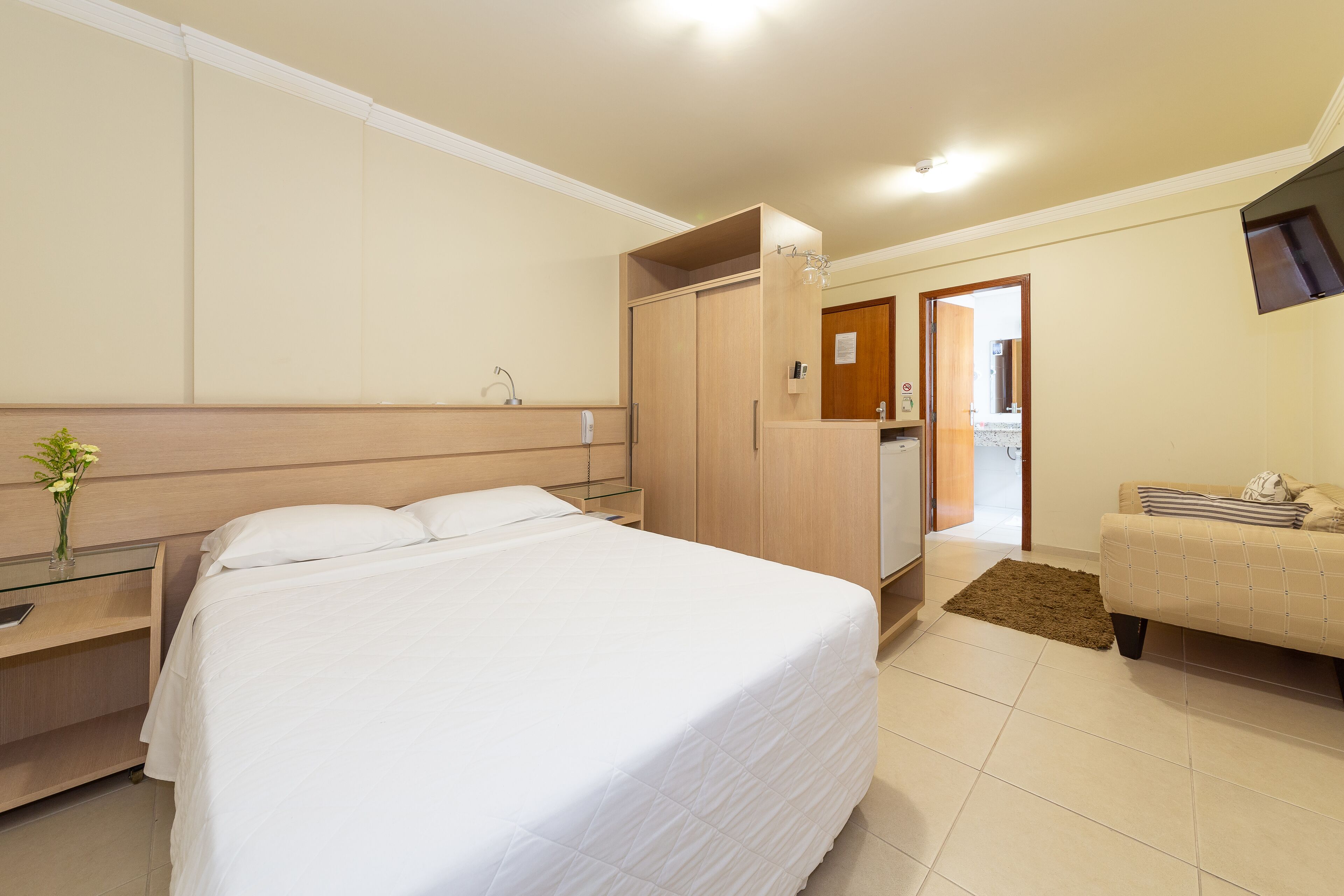 A hotel room with a large bed, a wooden wardrobe, a small desk with a lamp, and a sofa.