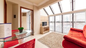 A modern living room with a glass table, red chairs, a red sofa, a TV on a wooden stand, and a large window with blinds. - SLIM São José Zion (Sao Jose)