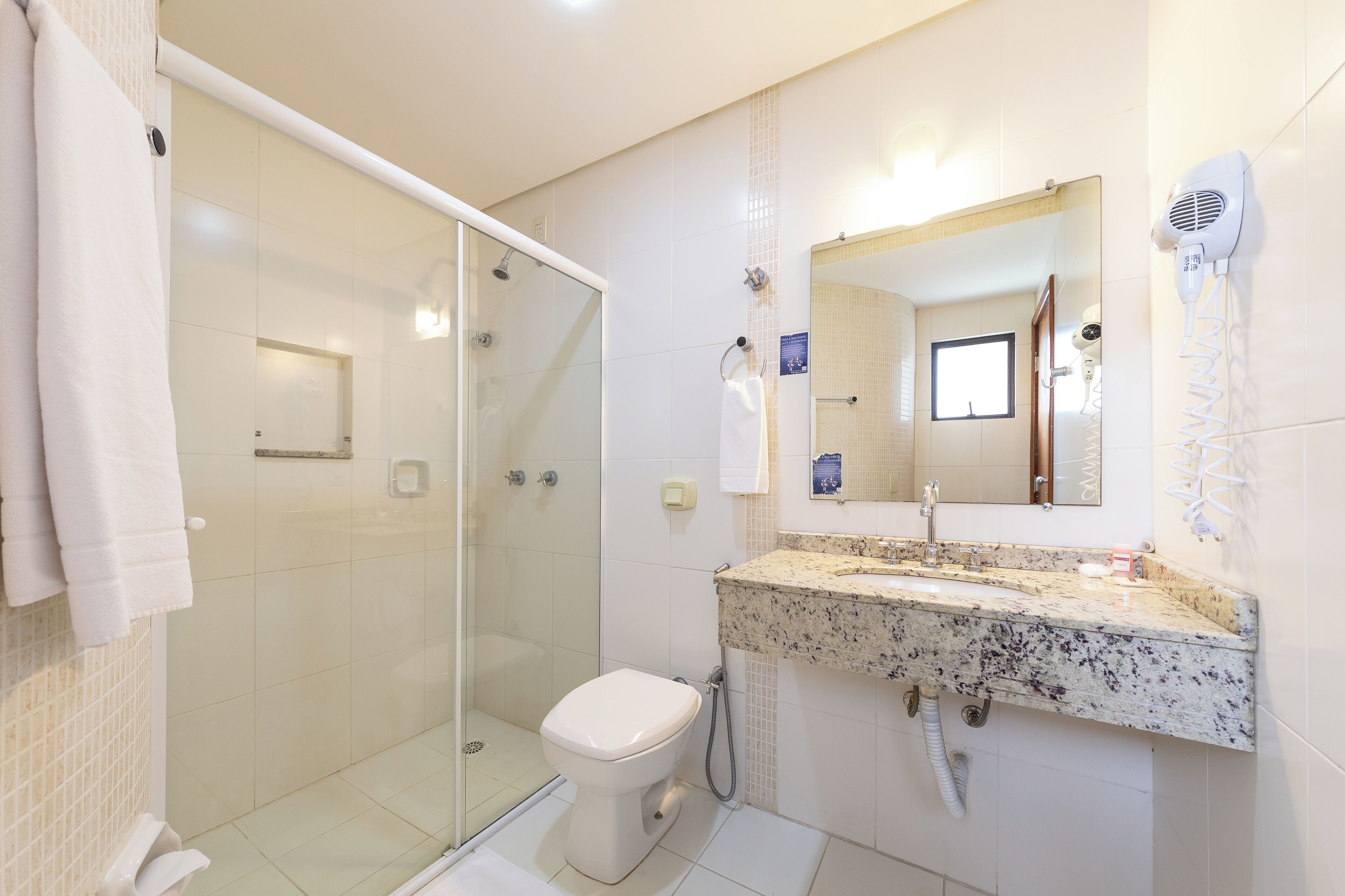 A bathroom with a glass shower enclosure, a white toilet, a granite countertop sink, and a hairdryer mounted on the wall.