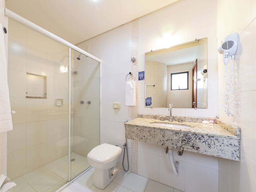 A bathroom with a glass shower enclosure, a white toilet, a granite countertop sink, and a hairdryer mounted on the wall.