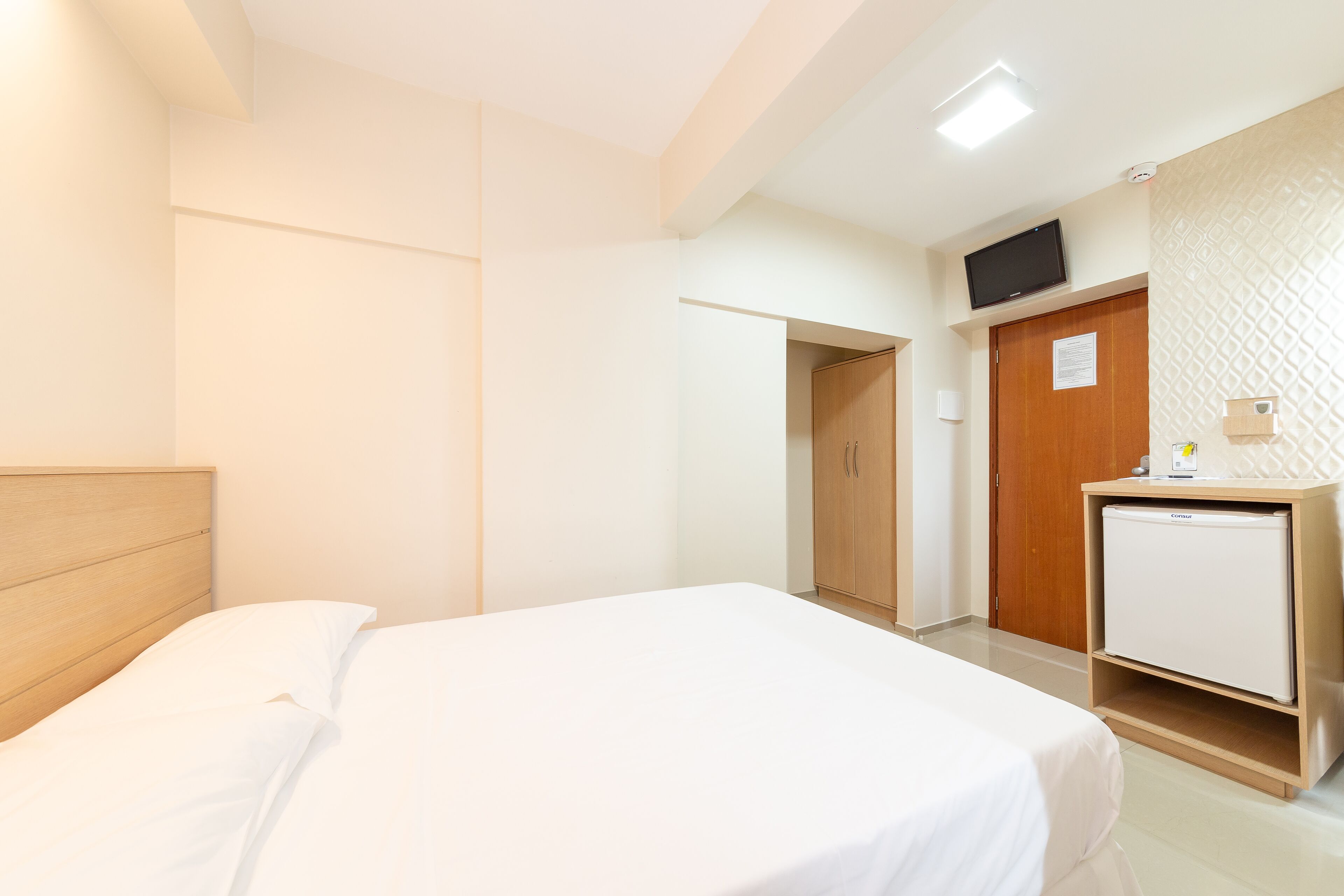 A hotel room with a bed, a wooden wardrobe, a small refrigerator, and a television on the wall.