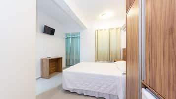 Un dormitorio con cama, televisión montada en la pared, armario de madera y ventana con cortinas.