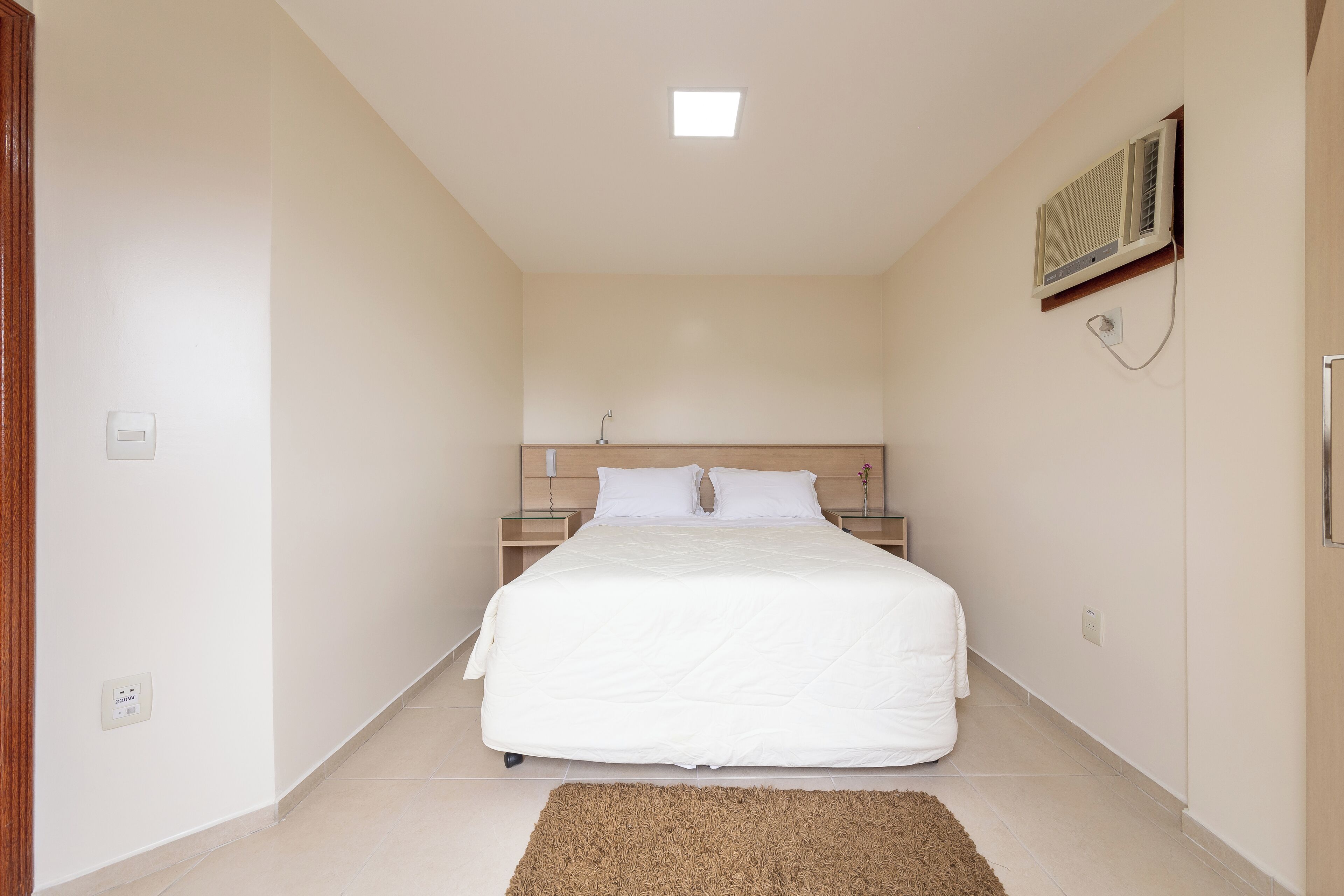 Une chambre à coucher comprenant un lit, des tables de chevet, un climatiseur fixé au mur et un luminaire au plafond.