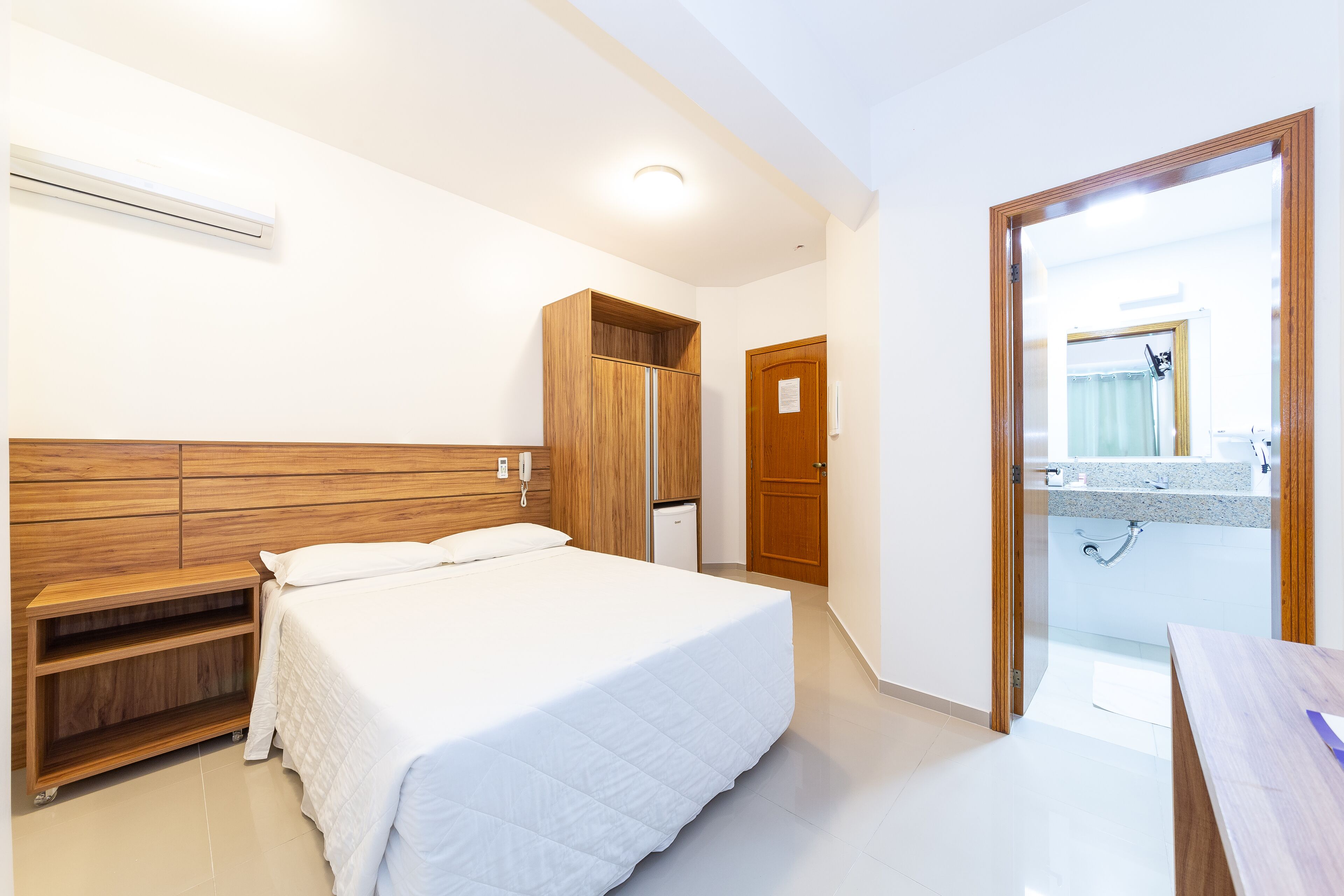 A bedroom with a bed, wooden wardrobe, built-in shelves, a small desk, and a bathroom visible through an open door.