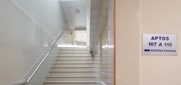 Escada com corrimão que leva a um andar superior. Uma placa indica 'APTOS 107 A 115' com uma seta apontando para a direita para 'ACESSO ESCADA'.