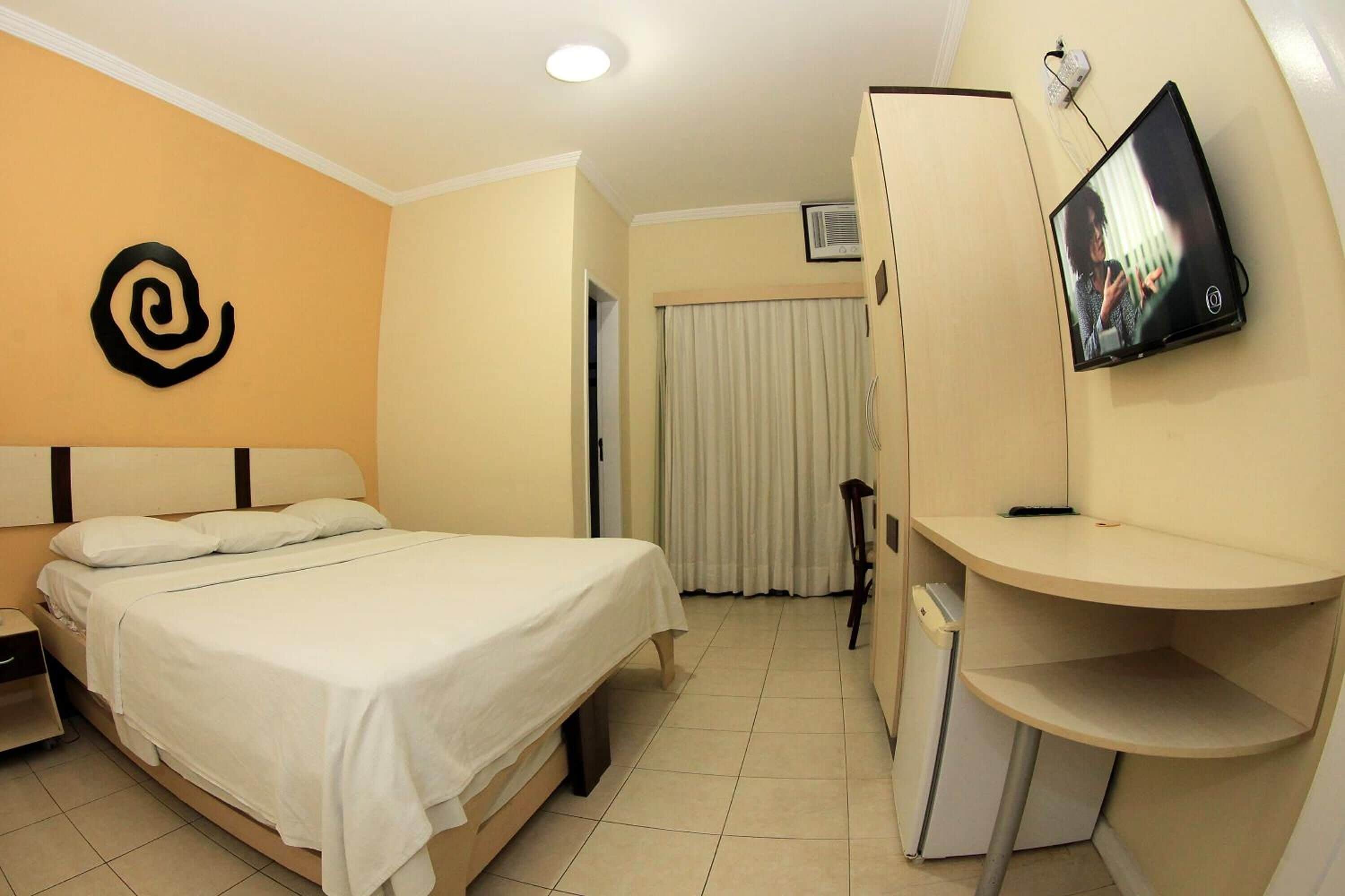 A hotel room with a bed, a desk, a television mounted on the wall, and a wardrobe.
