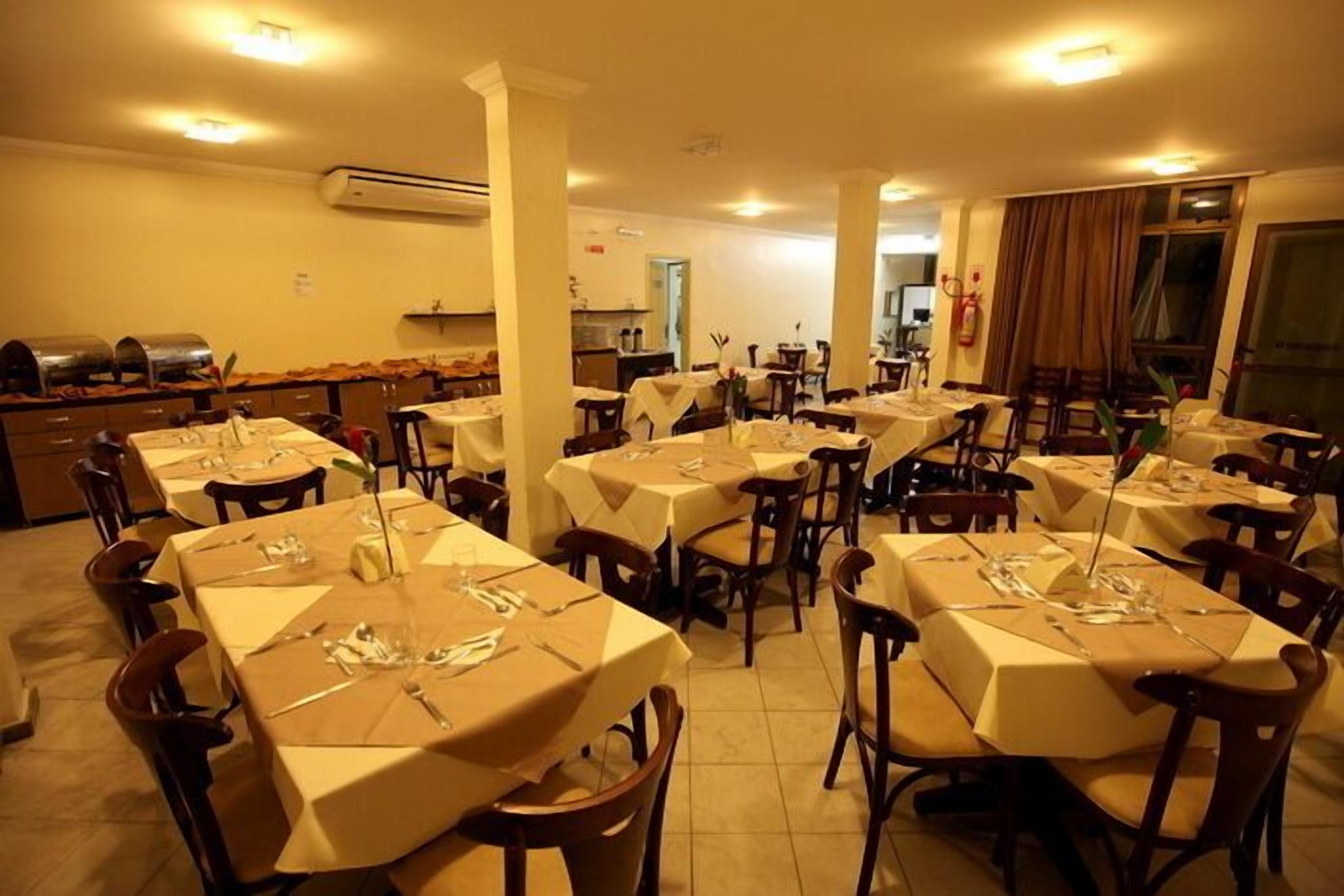 Une salle à manger avec plusieurs tables dressées, une cuisine à l’arrière-plan et une grande fenêtre avec des rideaux.