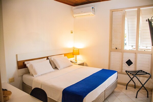 A hotel room with a bed, a nightstand with a lamp, an air conditioning unit, a window with shutters, and a small folding chair. - Hotel Terra do Sol (Aracaju)
