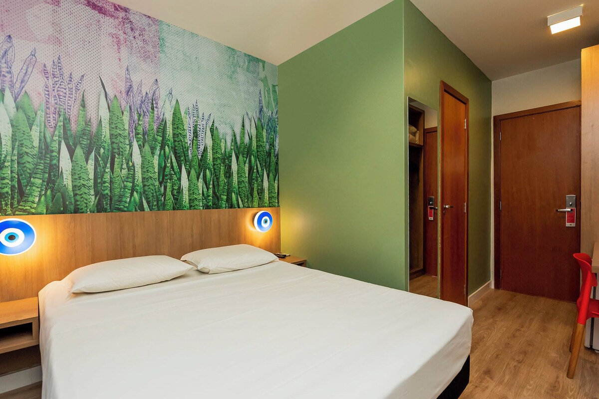 A hotel room with a bed, a wall-mounted artwork of plants, a wooden headboard, and a door with a red chair outside.