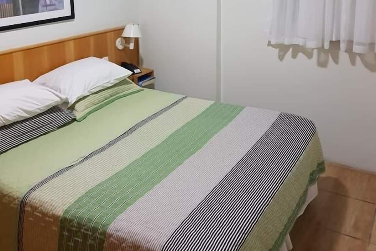 Quarto de hotel com uma cama, uma mesinha de cabeceira, uma luminária, um quadro na parede, uma janela com cortinas e uma luz no teto.