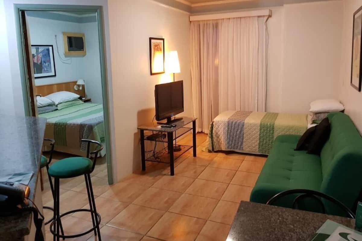 Quarto de hotel com cama, televisão em suporte, sofá verde, mesa pequena e área de bar.