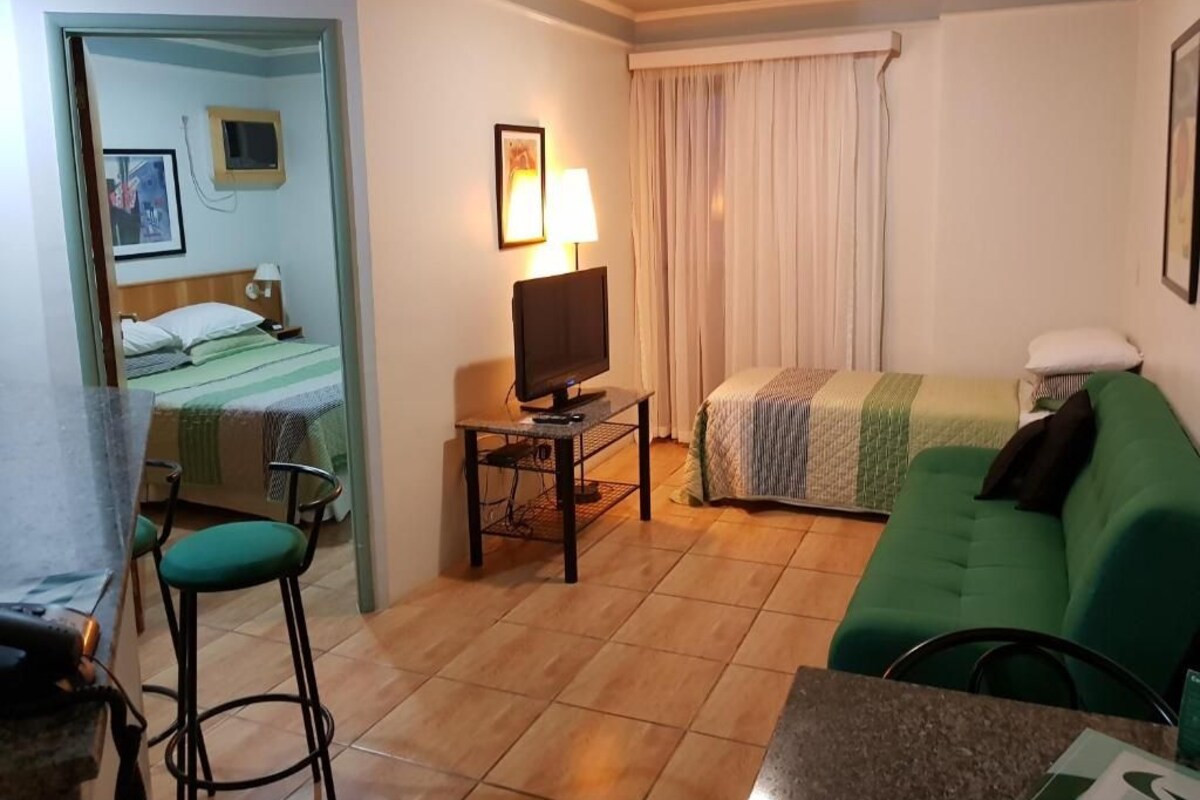A hotel room with a bed, a TV on a stand, a green sofa, a small table, and a bar area.