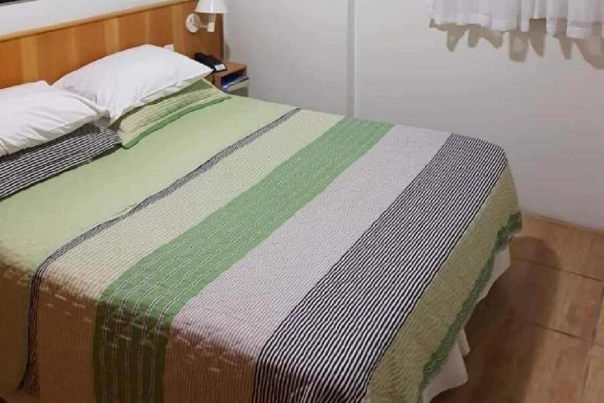 Quarto com cama, mesa de cabeceira, abajur, quadro na parede e janela com cortinas.