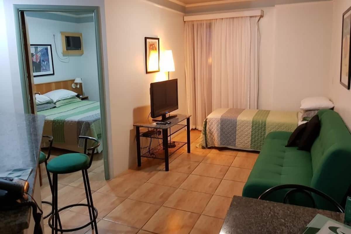 Quarto de hotel com cama, televisão sobre uma mesinha, sofá verde e uma cadeira.