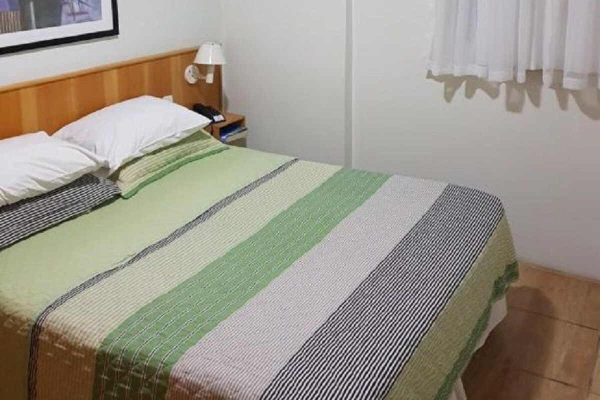 Un dormitorio con cama, mesita de noche, lámpara, un cuadro en la pared y ventana con cortinas.