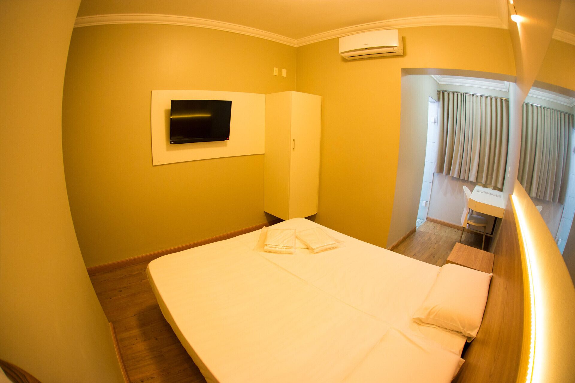 Quarto de hotel com cama, televisão na parede, ar condicionado, escrivaninha com cadeira e janela com cortinas.