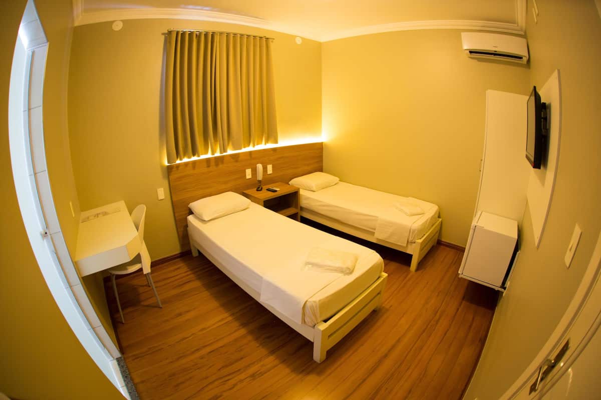 Ein Hotelzimmer mit zwei Einzelbetten, einem Schreibtisch, einem Stuhl, einem Nachttisch, einer Klimaanlage und einem an der Wand befestigten Fernseher.