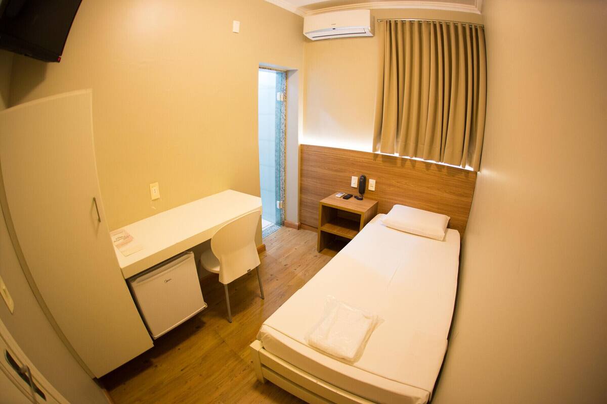 Ein kleines Hotelzimmer mit einem Bett, einem Schreibtisch, einem kleinen Kühlschrank, einem Nachttisch mit Telefon und einer Klimaanlage.