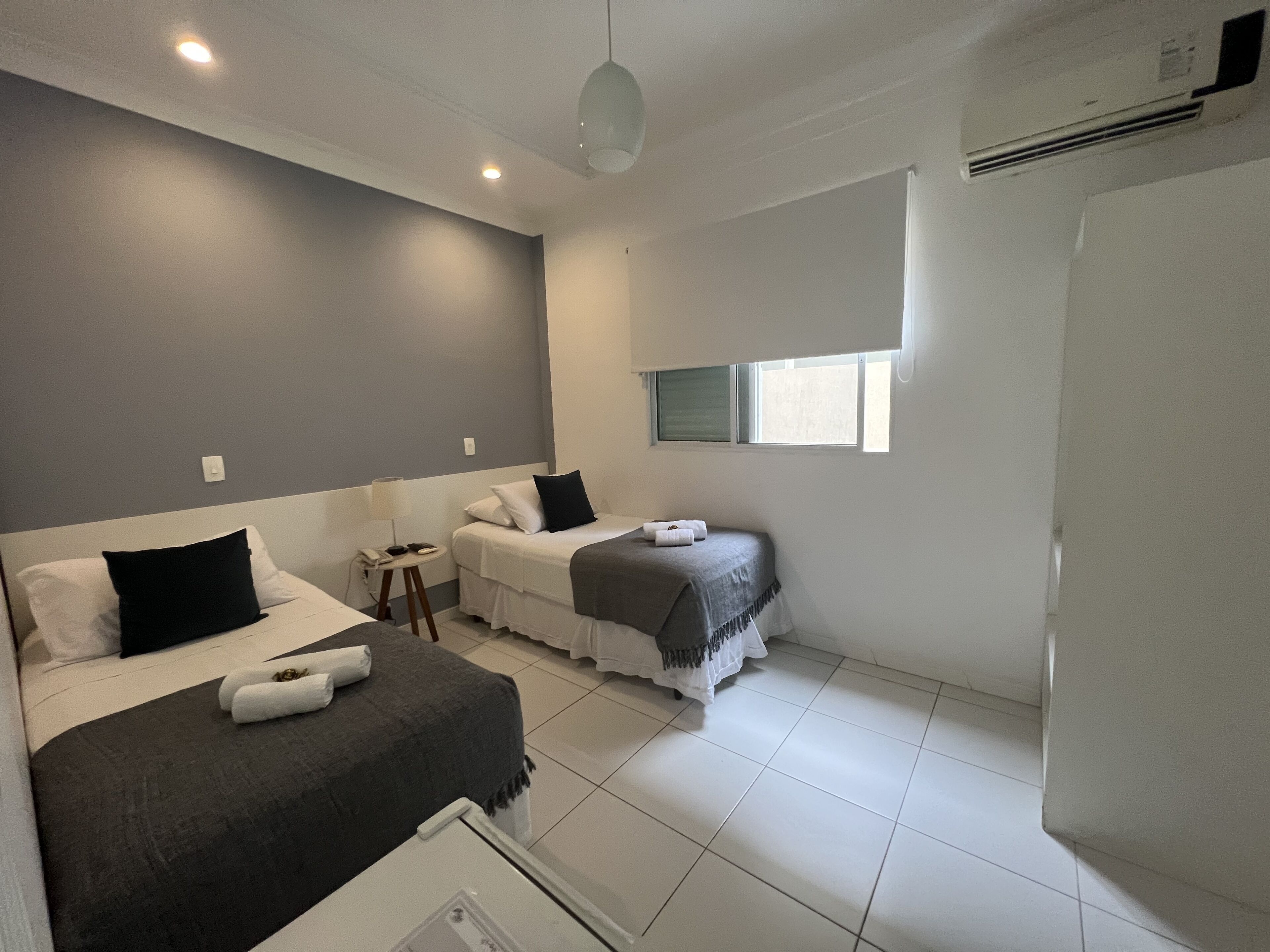 Quarto com duas camas, uma mesinha, uma janela e um aparelho de ar condicionado.
