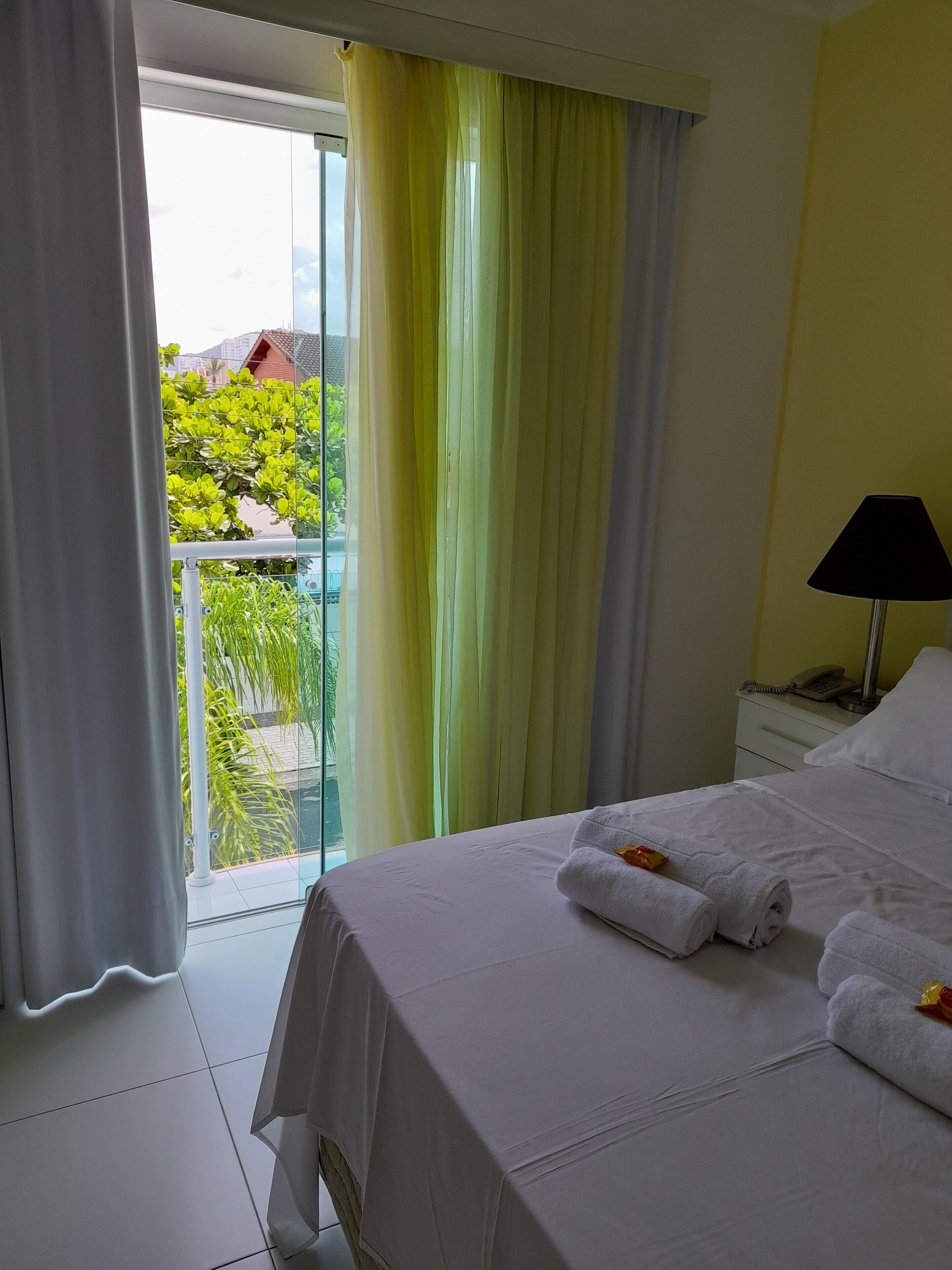 A hotel room with a bed, a nightstand with a lamp, and a view of greenery outside.