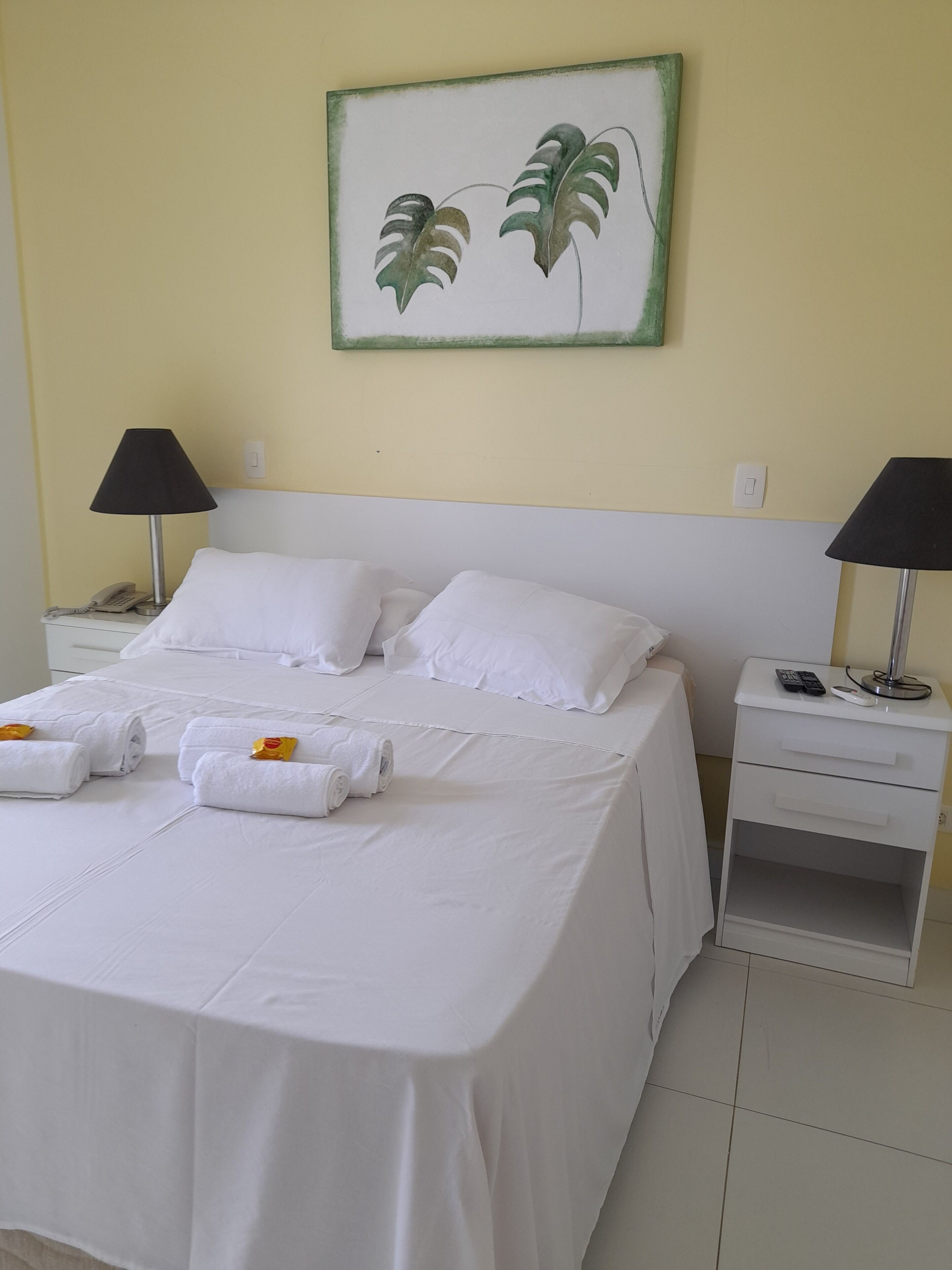 Cama bem arrumada com lençóis e almofadas brancas, duas mesas de cabeceira com luminárias, um telefone e um quadro de folhas na parede.