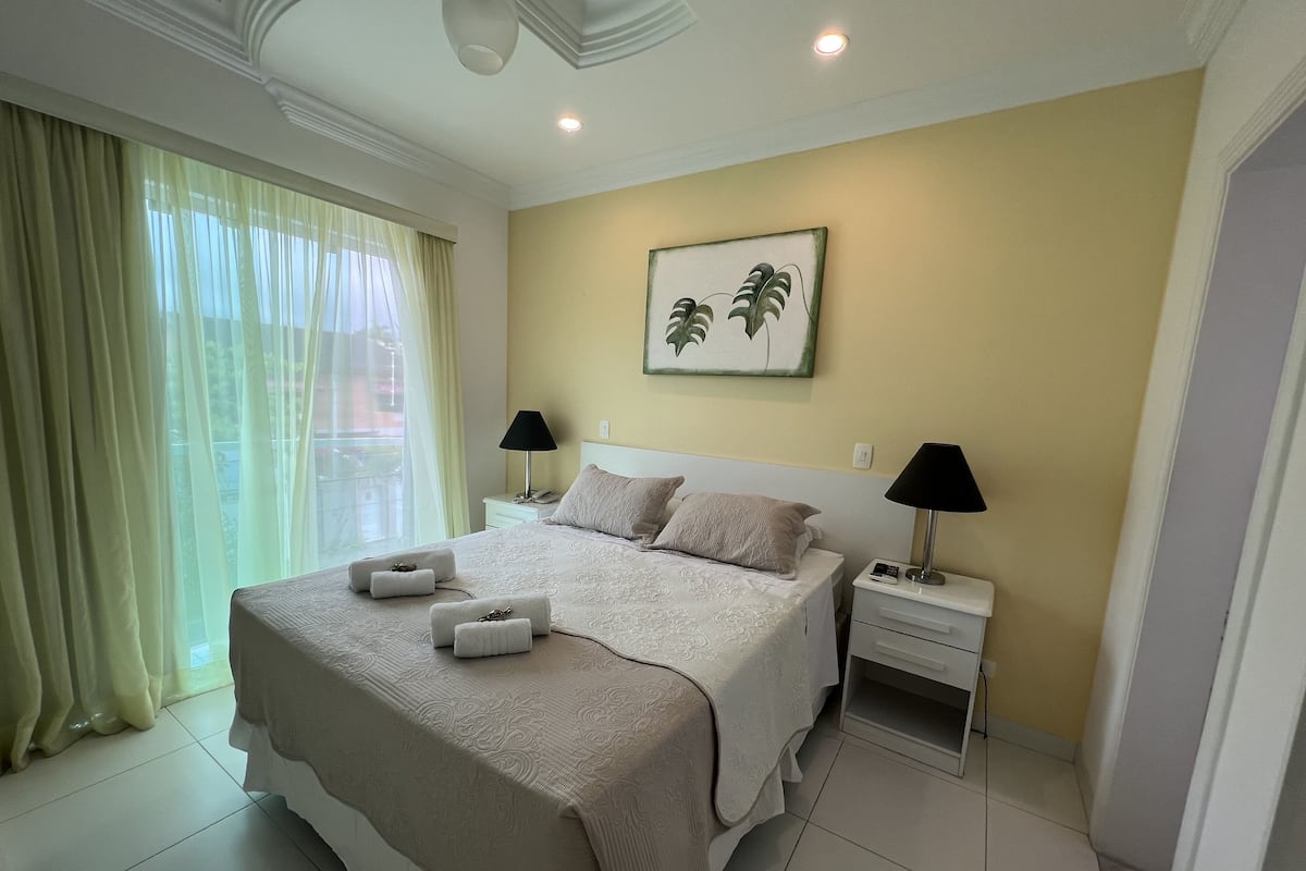 A neatly arranged bedroom with a large bed, two bedside tables, a wall-mounted painting, and a window with sheer curtains.