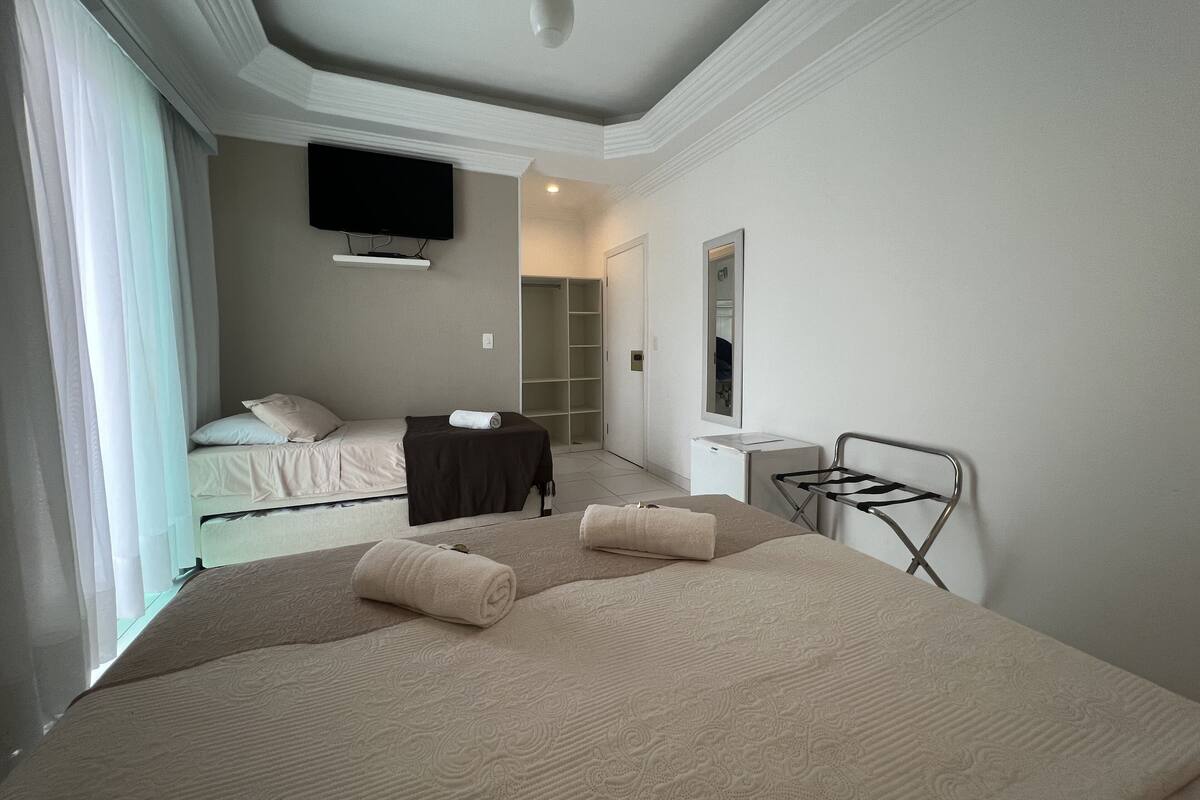 A modern hotel room with a large bed, a TV mounted on the wall, a mirror, and a small desk with a lamp.