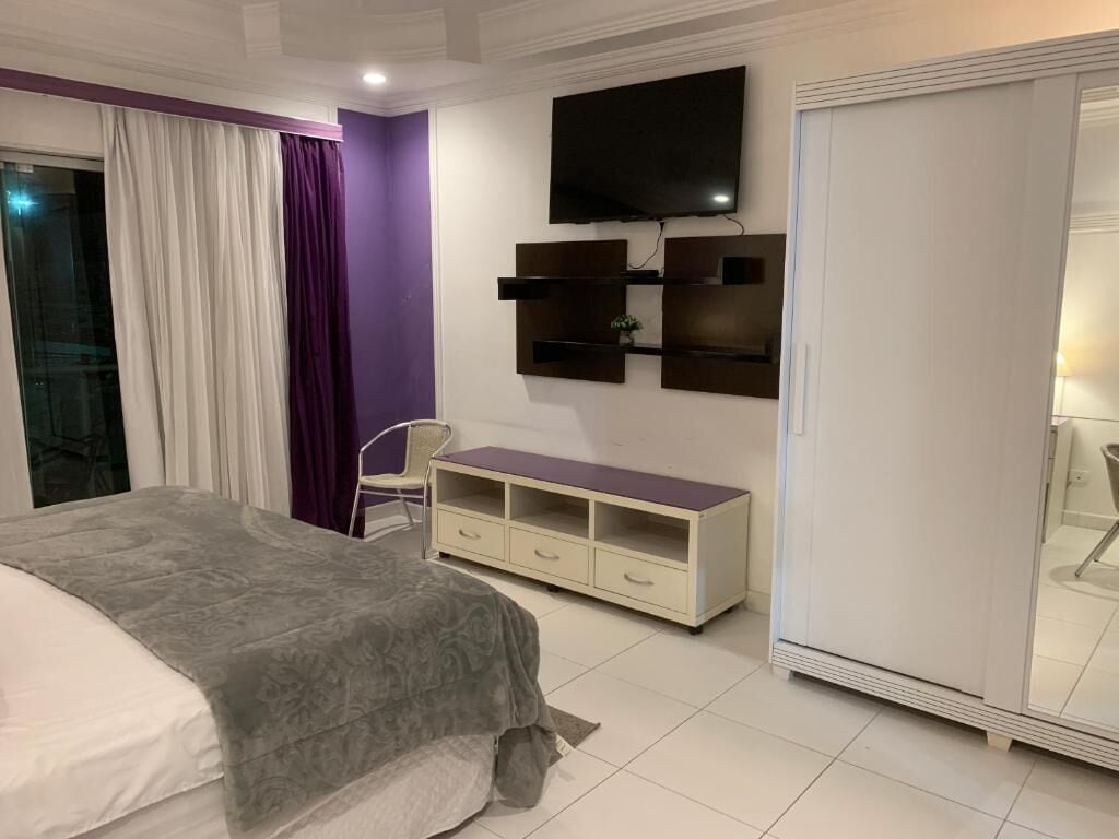 Quarto com cama, televisão na parede, guarda-roupa e um pequeno armário.