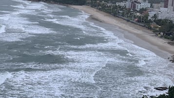 Vista costeira com praia, ondas do oceano e uma colina arborizada ao fundo.