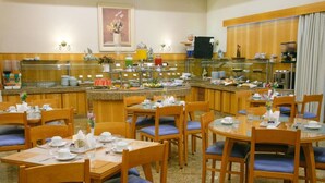 Un comedor de hotel con varias mesas y sillas, un mostrador de buffet con diversas opciones de comida y un ventanal con cortinas.