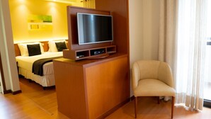 Habitación de hotel con cama, televisor sobre un soporte de madera, una silla y un ventanal con cortinas.