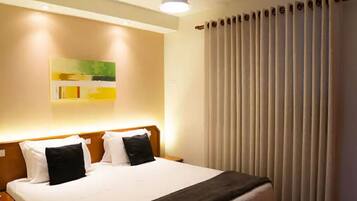 A hotel room with a bed, a ceiling fan, a wall-mounted light, and a window with vertical blinds.