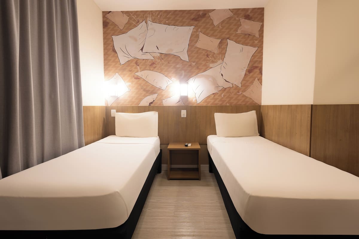 Quarto de hotel com duas camas de solteiro, cabeceira de madeira, mesinha de cabeceira com controle remoto e parede com desenho estampado.
