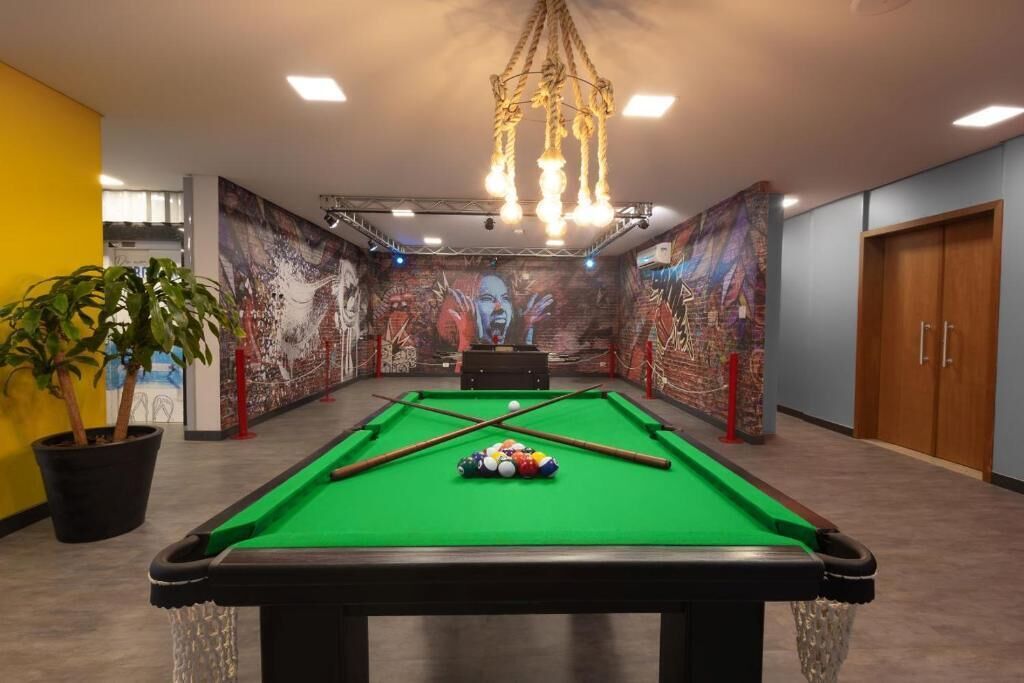 Uma mesa de bilhar com bolas no centro de um salão com painel nas paredes e um candelabro acima.