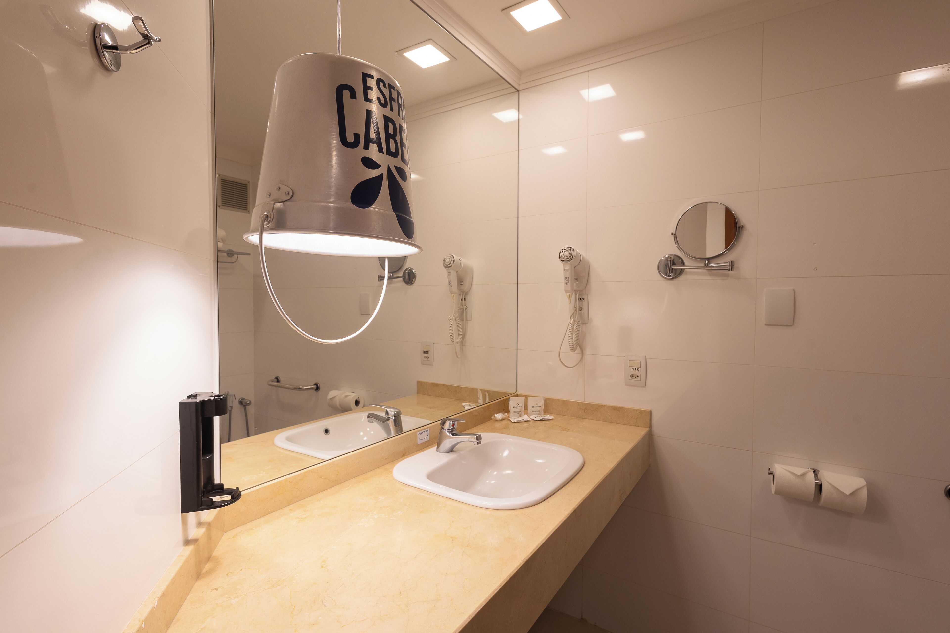 Une salle de bain moderne avec un comptoir de couleur beige, deux lavabos, un grand miroir, un porte-serviettes et un support à papier toilette.