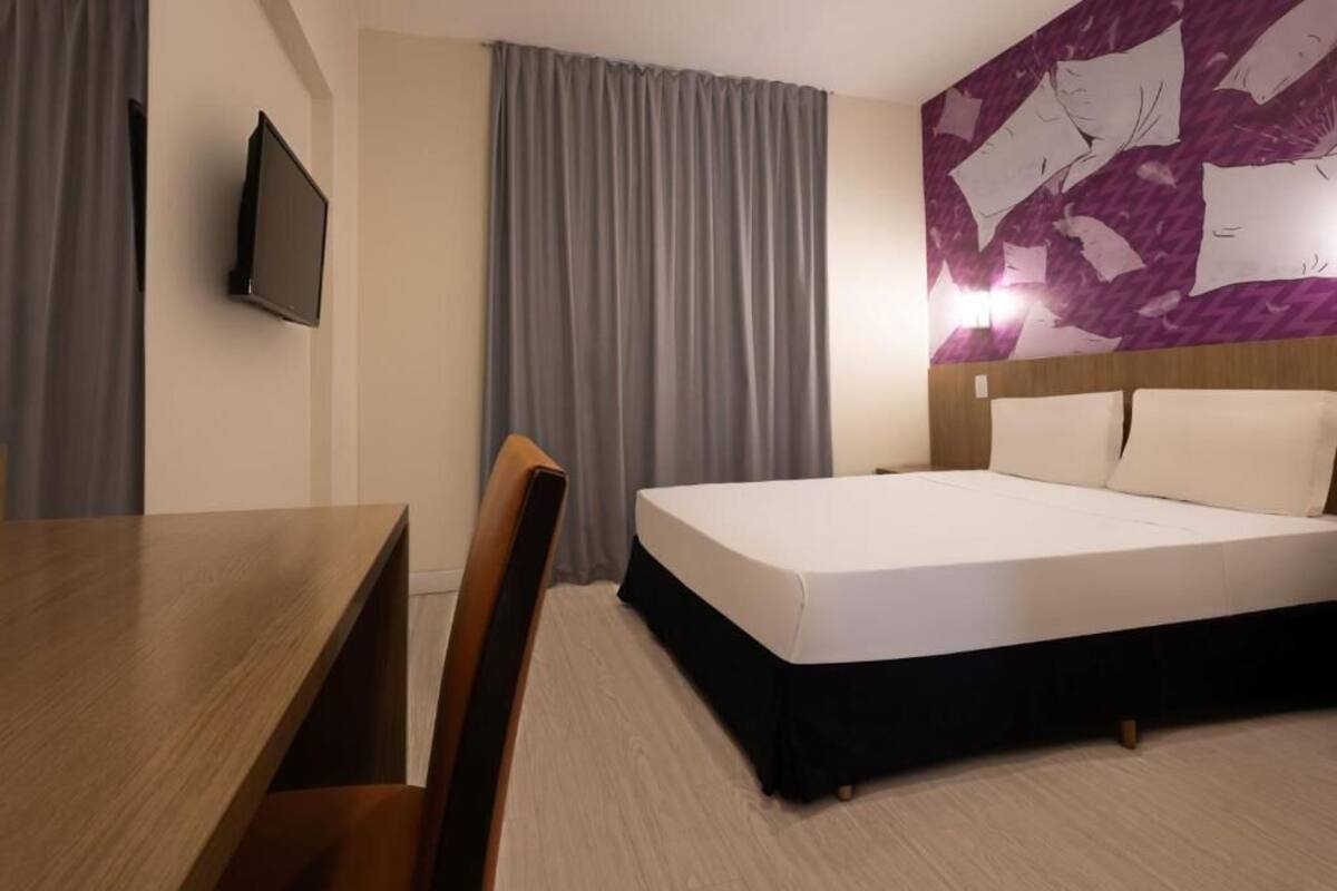 Quarto de hotel com uma cama, uma escrivaninha e uma televisão.