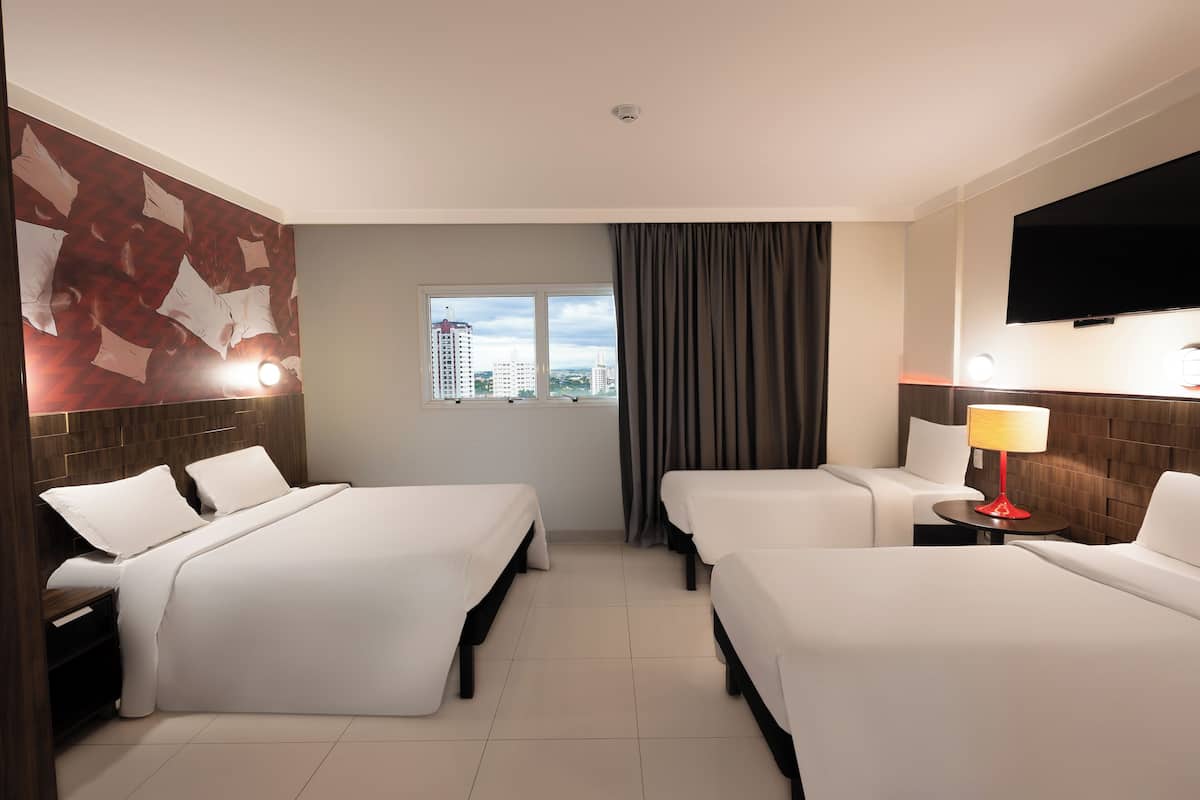 Quarto de hotel com duas camas, televisão, janela com cortinas e vista para edifícios.
