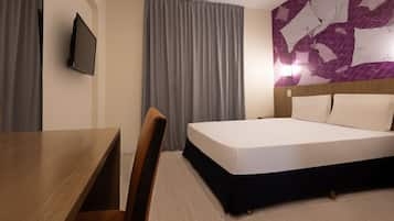 A hotel room with a bed, a desk, a chair, a TV, and a wall with a geometric pattern.