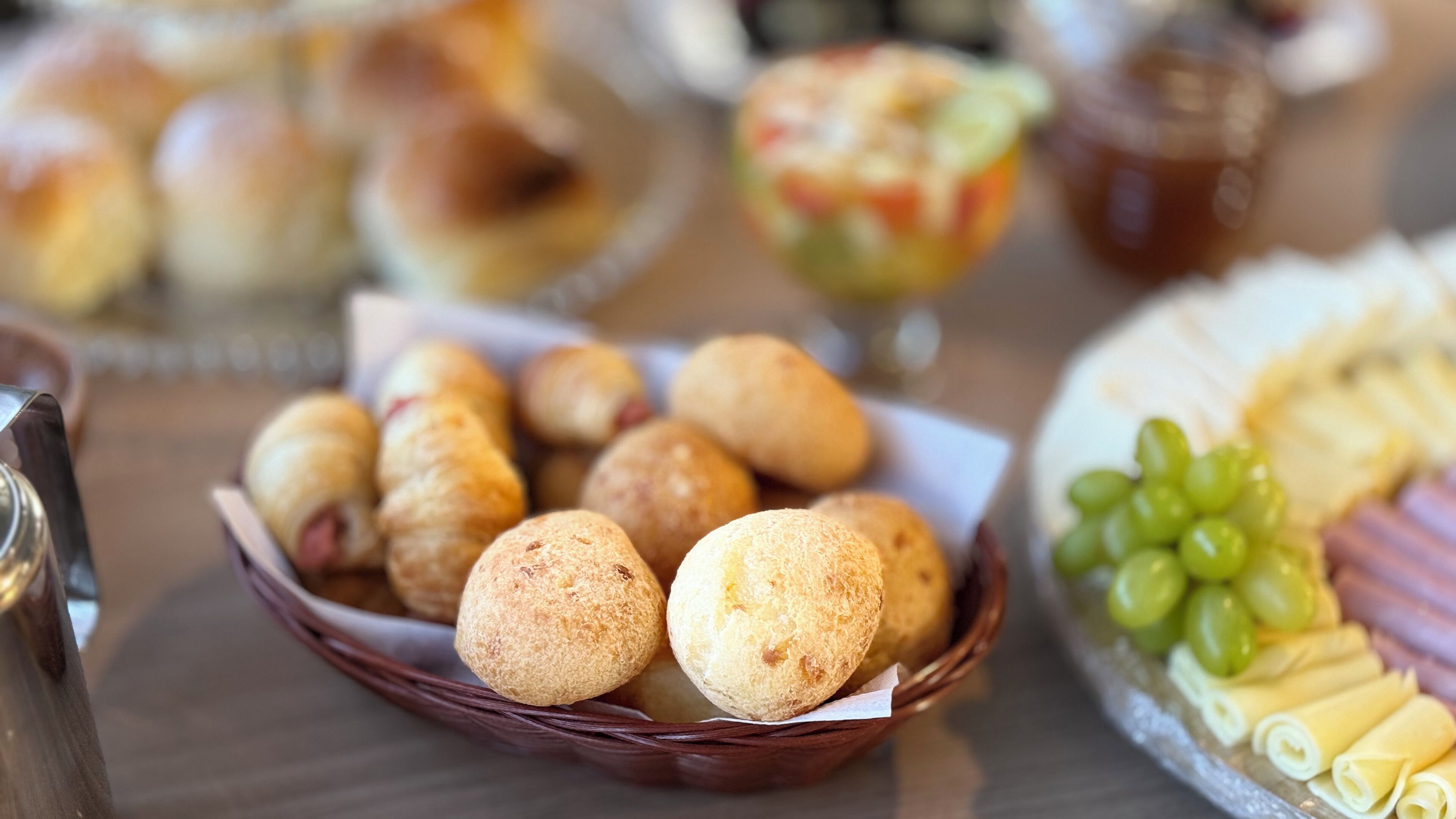 A table with a basket of round pastries, a plate of sliced cheese and grapes, and a glass of beverage.