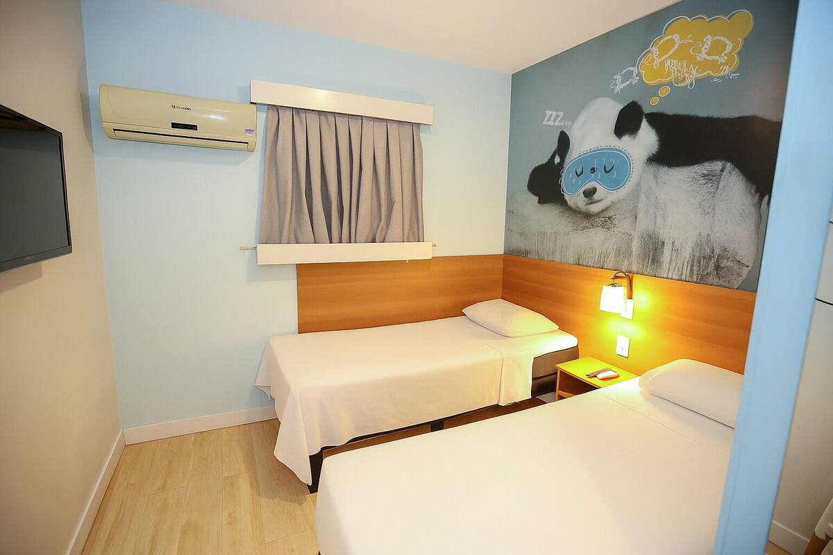 A hotel room with two single beds, a large wall mural, an air conditioning unit, and a bedside table with a lamp.