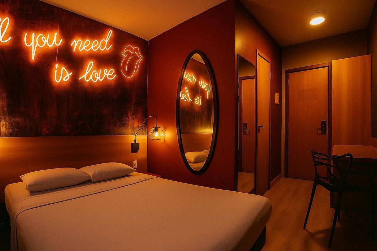 A hotel room with a bed, a neon sign, a mirror, and a door.