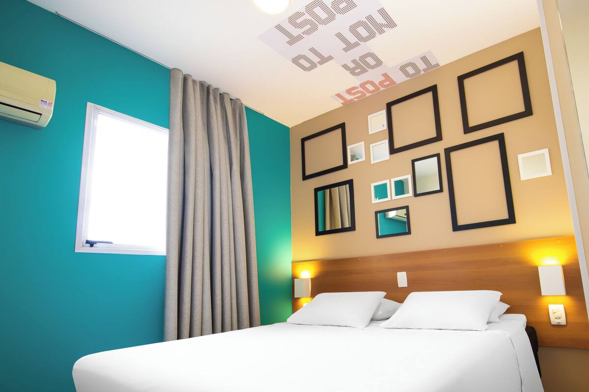 A hotel room with a bed, curtains, a window, wall-mounted lights, and framed pictures.