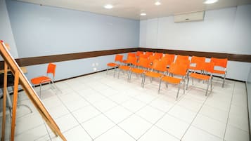 Sala de conferência com cadeiras laranja dispostas em fileiras, uma escada de madeira e piso revestido de azulejos brancos.