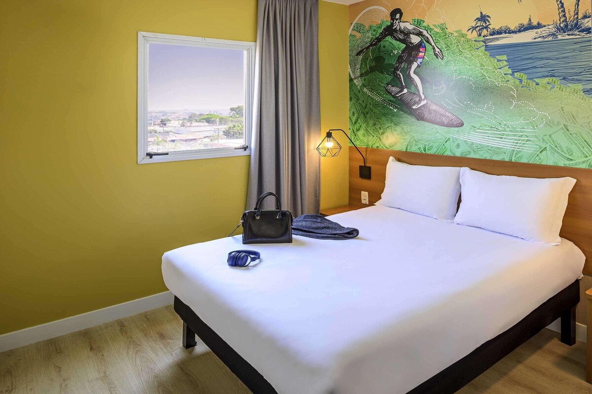 A hotel room with a bed, a window, a painting, and a lamp.