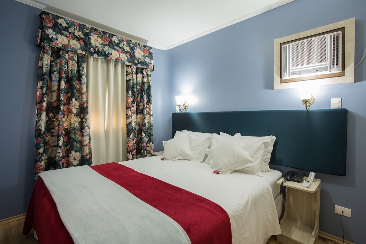 A hotel room with a bed, floral curtains, a nightstand with a phone, and a wall-mounted air conditioner.