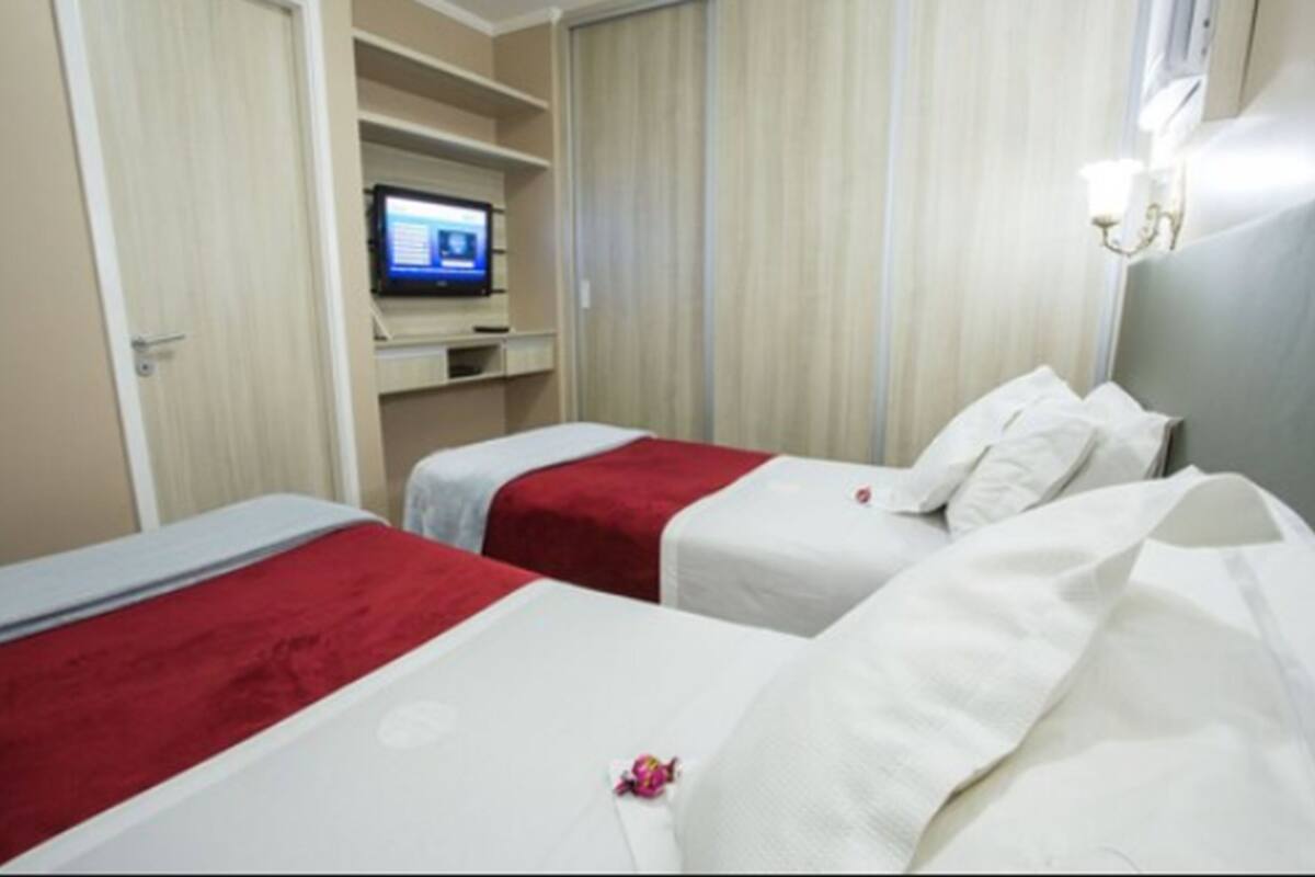 A hotel room with two beds, each with a red and white bedspread. There is a television on a shelf above the beds, and a window with curtains.