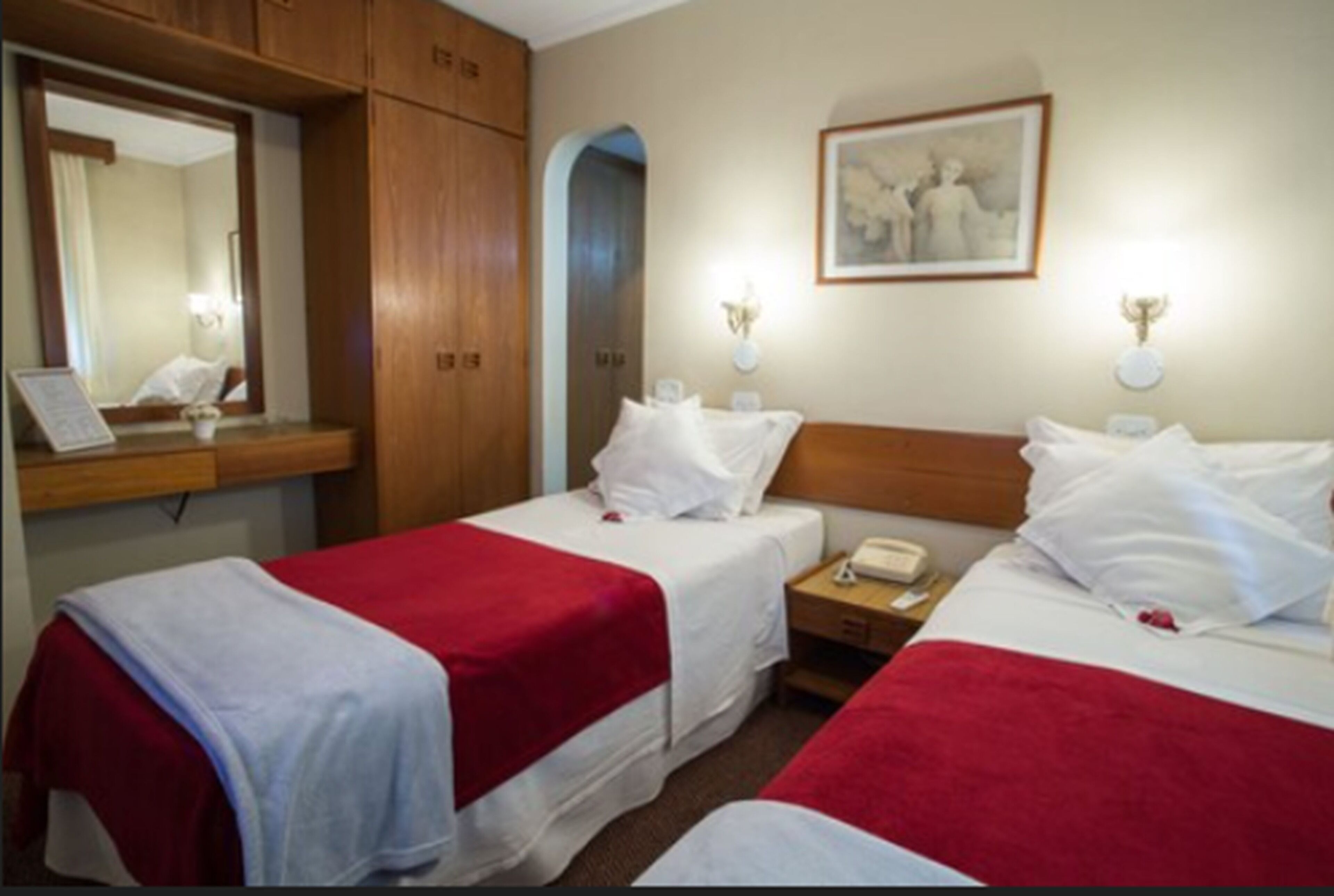 Une chambre d’hôtel avec deux lits simples, une armoire en bois, un miroir, un tableau encadré au mur et des lampes de chevet.
